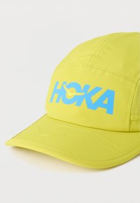 Φωτεινό κίτρινο καπέλο από ελαφρύ ύφασμα με μπλε λογότυπο "HOKA", κυρτό γείσο και εμφανείς ραφές για αντοχή.