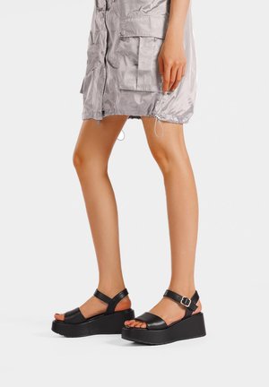 Gambe con sandali neri con zeppa e cinturini alla caviglia, abbinati a una gonna corta grigio chiaro lucida con grandi tasche frontali.