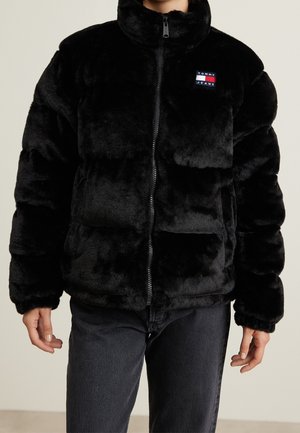 Persoon draagt een zwart pluche ritssluiting jas met een Tommy Jeans-logo op de borst en donkergrijze broek, staand tegen een neutrale achtergrond.