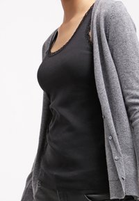 Kvinna som bär en svart linne med spetsdetaljer under en grå knapplåst cardigan, som visar överkropp och del av armar mot en vit bakgrund.