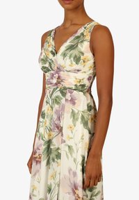 Blumen Kleid mit V-Ausschnitt, gefälteltem Oberteil und weitem Rock. Der Stoff zeigt Pastellfarben mit grünen Blättern, gelben und lila Blumen.