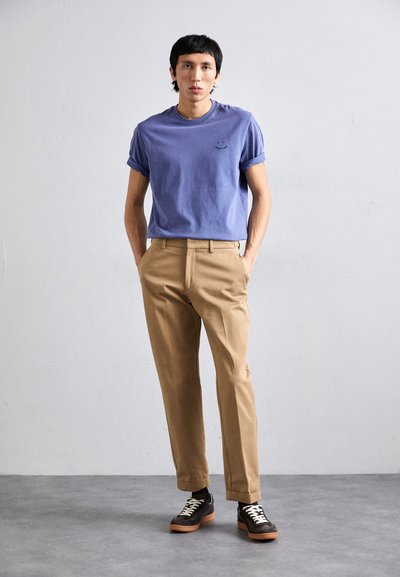 Ανοιχτό μοβ βαμβακερό μπλουζάκι με ρολάκια στα μανίκια, καφέ κομμένα παντελόνια και σκούρα αθλητικά παπούτσια με λευκούς κορδόνια. Απλή, μοντέρνα σιλουέτα.