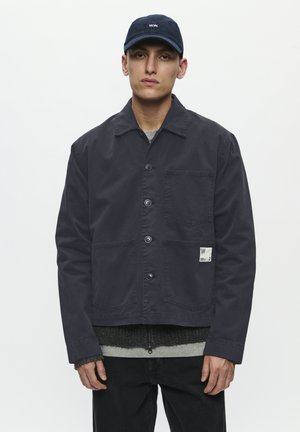 Dark navy jacket made of sturdy cotton with a classic collar, front buttons, two patch pockets, and a small brand label. Textured grey sweater beneath.