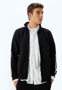 Sudadera negra con cremallera y rayas laterales blancas, con puños y dobladillo acanalados, hecha de una tela suave, llevada sobre una camisa blanca de botones.