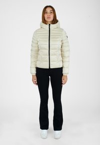 Giacca imbottita crema con cappuccio, caratterizzata da una cerniera frontale e polsini elasticizzati. Indossata sopra pantaloni neri e sneakers bianche. Sfondo bianco.