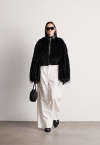 Casaco de pelúcia preta com mangas oversized, combinado com calças brancas de perna larga e botins pretos de bico fino. Acessório: pequena bolsa preta.