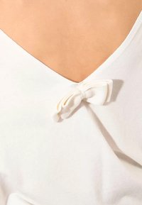 Blusa bianca con scollo a V e un piccolo dettaglio a fiocco in tessuto sul retro. Tessuto morbido e design minimalista.