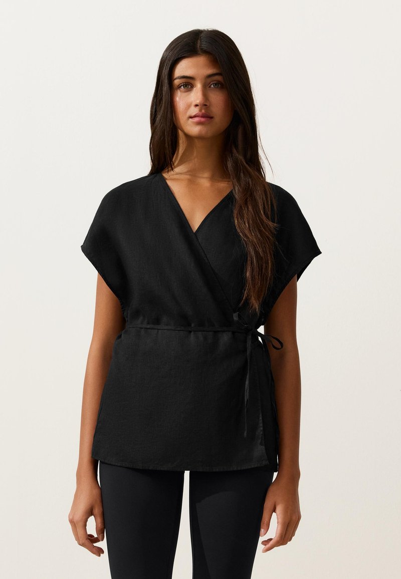 Femme aux longs cheveux noirs portant un haut noir à manches courtes croisé et noué à la taille, associé à un pantalon noir, debout devant un fond uni.