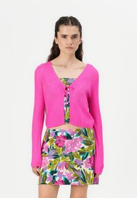 Cardigan corto di un rosa acceso con texture a coste e maniche lunghe, dotato di chiusura anteriore a fiocco. Abbinato a una gonna a stampa floreale vivace.