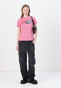Camiseta de algodón rosa con logotipo negro de Diesel, combinada con pantalones cargo negros de corte holgado y zapatos de plataforma negros gruesos.