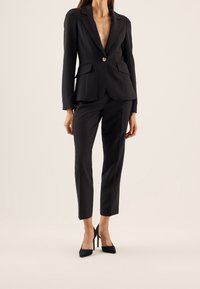 Black blazer with button closure and sharp lapels, paired with tailored black trousers and pointed black heels. Smooth fabric texture.