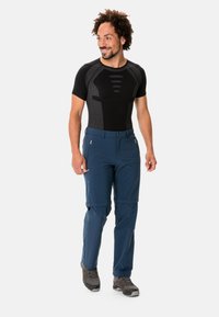 Camicia nera aderente da uomo con accenti testurizzati, abbinata a pantaloni blu navy con zip. L'outfit è completato da scarponi da trekking grigi.