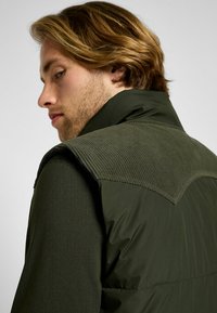 Gilet verde con una zona delle spalle in corduroy testurizzato, caratterizzato da una parte inferiore liscia e imbottita. Il tessuto sembra leggero e resistente.