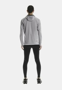 Veste de sport à capuche gris clair avec des accents noirs, associée à des leggings noirs ajustés et des chaussures robustes, vue de dos.