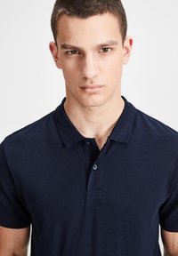 Marineblaue Baumwollpolohemd mit Kragen, drei Knöpfen am Hals und einem kleinen Logo-Detail auf der Brust. Kurze Ärmel, schlichte Textur.