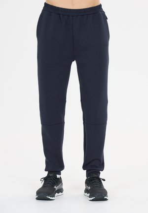 Mann, der schwarze Sporthosen mit elastischem Bund und schwarze Laufschuhe trägt, steht vor weißem Hintergrund.