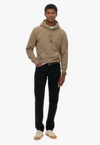 Beige hoodie met trekkoorden en een voorzak, gecombineerd met zwarte jeans en beige sneakers. Minimalistisch ontwerp, zachte textuur.