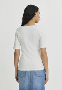 Femme aux longs cheveux foncés portant un haut blanc côtelé à manches courtes et une jupe en denim bleue, vue de dos sur un fond uni.