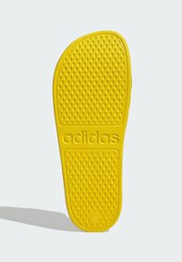 Suela de goma amarilla con una superficie texturizada que presenta pequeños patrones circulares y la marca "adidas" en relieve. Forma redondeada con un ligero estrechamiento.