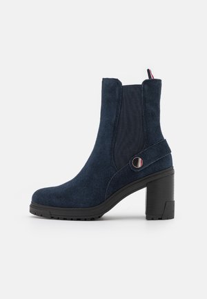 Donkerblauwe suède enkellaars met elastische zijkant, decoratieve riem met metalen knoop en stevige zwarte blokhak.