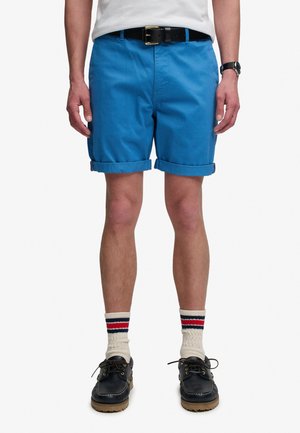 Mężczyzna w jasnoniebieskich krótkich spodenkach z podwiniętymi krawędziami, czarnym paskiem, białych skarpetkach z czerwonymi i czarnymi paskami, czarnych sznurowanych butach i zegarku na lewej ręce.