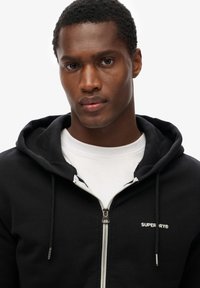 Felpa con zip nera con il logo bianco "SUPERDRY" sul lato sinistro del petto, dotata di cappuccio con cordino e una trama di tessuto liscia.