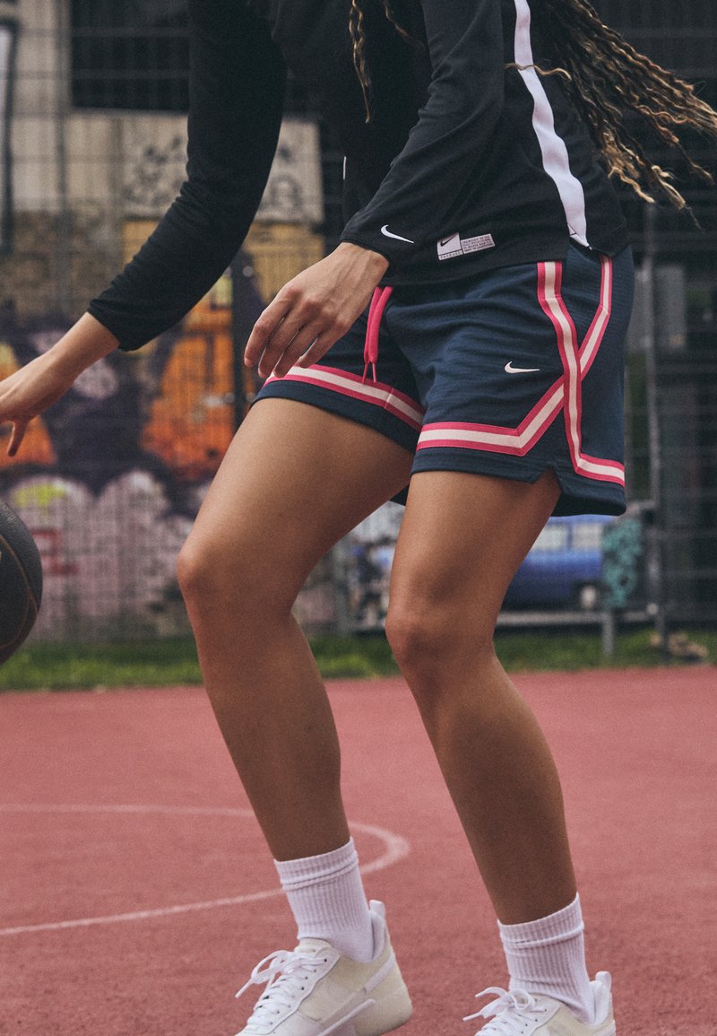 Tumesinised sportlikud lühikesed püksid roosa ja valge aktsentidega, koos pika musta varrukaga särgiga ja valgete tossudega. Korvpall käes väljakul.