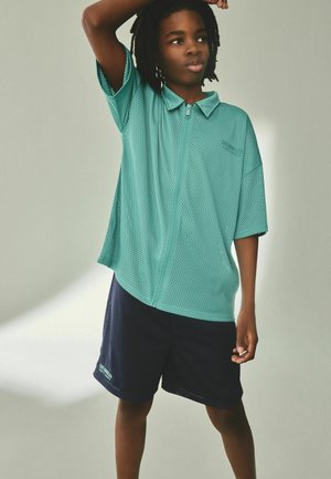 Ung person med korte dreadlocks iført en turkis lynlåstrøje i mesh og marineblå mesh-shorts, stående med den ene arm hævet.