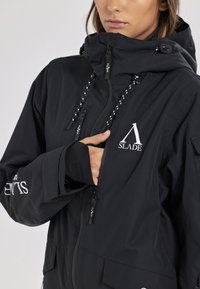 Giacca nera impermeabile con colletto alto, tasche con zip, cappuccio regolabile con lacci e un logo prominente sul davanti.