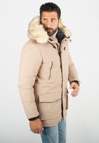 Parka beige avec une capuche en fausse fourrure, des toggle à l'avant, des poches zippées et une coupe décontractée. Tissu texturé avec des accents noirs contrastants.