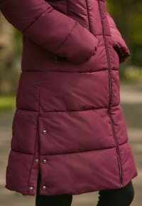 Bordo puffer kaput s kvadratnim dizajnom, s džepovima na zatvarač i srebrnim dugmićima na rubu, nosi se s crnim tajicama.