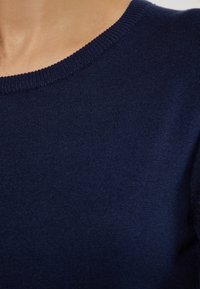 Námorně modrý pletený svetr s vroubkovaným kulatým výstřihem, detailní pohled, který ukazuje hladkou texturu a jemné detaily látky.