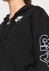 Černá mikina na zip s designem mesh na ramenech, s bílým logem Nike a vyšitými detaily na rukávech. Měkká tkanina s kapsami.