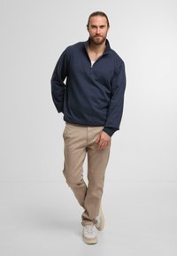 Granatowa bluza z zamkiem i wysokim kołnierzem, noszona na białej koszuli, zestawiona z beżowymi spodniami i białymi sneakerami. Prosty, luźny krój.