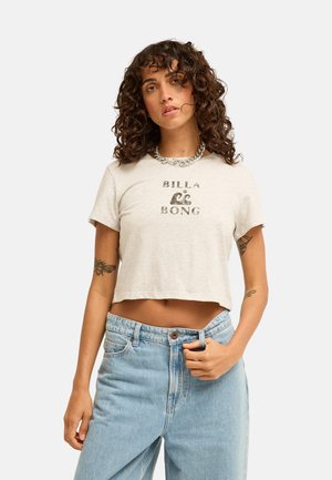 Kobieta z kręconymi włosami ubrana w beżowy krótki t-shirt Billabong, niebieskie jeansy, naszyjnik z łańcuszka oraz prezentująca tatuaże na obu ramionach.