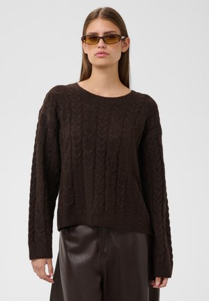Femme portant un pull en maille torsadée marron, un pantalon en cuir foncé et de petites lunettes de soleil teintées ambre sur un fond clair uni.