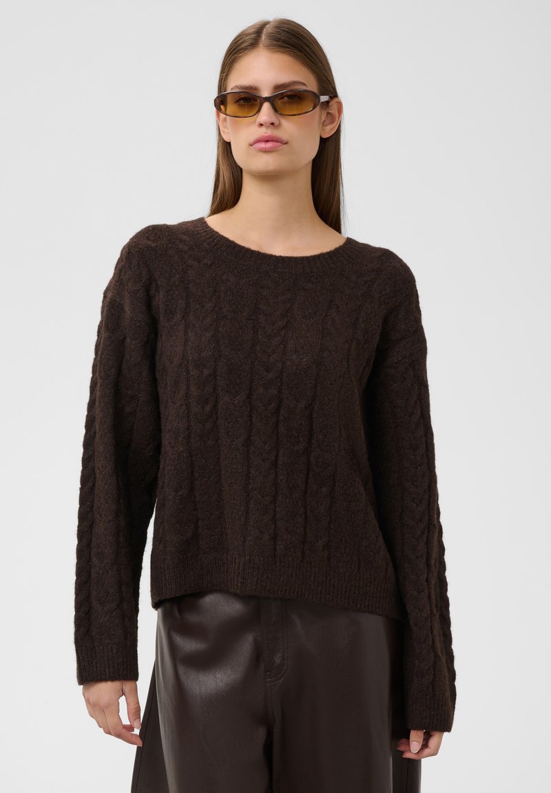 Femme portant un pull en maille torsadée marron, un pantalon en cuir foncé et de petites lunettes de soleil teintées ambre sur un fond clair uni.