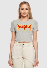 Pelēks īsa krekls ar "BADGIRLS" logo, kas izceltas bolder sarkanā un dzeltenā krāsā ar asiem, leņķainiem akcentiem. Gluda kokvilnas audums.