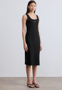 Μαύρο ριμπ φόρεμα με στενή σιλουέτα, με στρογγυλή λαιμόκοψη και λεπτομέρεια με λογότυπο στο στήθος. Το μοντέλο φοράει μαύρα σανδάλια τύπου slide.