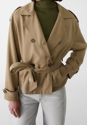 Vrouw draagt een beige dubbelgeknoopte jas met riem over een olijfgroene geribde coltrui en lichtgrijze broek.