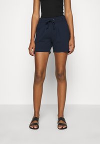 Donna che indossa pantaloni corti blu navy con cordoncino, top senza maniche nero e sandali neri con suola piatta, in piedi di fronte a uno sfondo bianco.