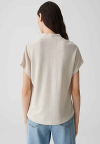 Top beige a maniche corte realizzato in tessuto texturizzato, caratterizzato da una discreta lucentezza, vestibilità ampia e orlo arrotondato, abbinato a jeans azzurri chiari.