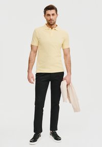 Żółta koszulka polo z kołnierzykiem, czarne spodnie slim-fit i czarne trampki. Osoba trzyma jasnobeżową kurtkę. Gładkie tło.