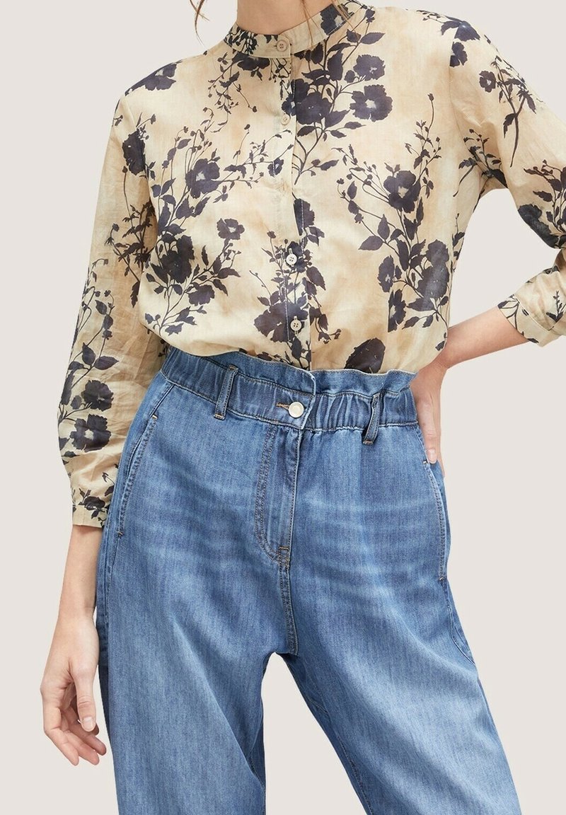 Blouse beige à manches longues avec motifs floraux noirs, associée à un jean bleu clair taille haute avec une ceinture élastique.