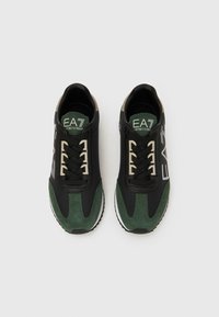 Fekete és zöld sportcipő, sima és velúr textúrák keverékével, fehér talppal és fűzővel, az „EA7” márkajelzéssel a nyelvén.