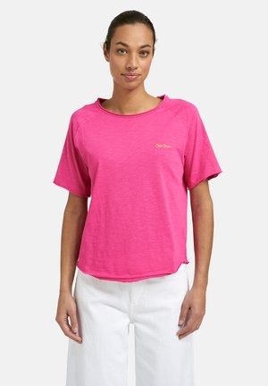 Frau trägt ein knallpinkes Kurzarmshirt mit kleinem gelben "Cape Town" Schriftzug auf der linken Brust, kombiniert mit weißen Hosen, stehend vor weißem Hintergrund.