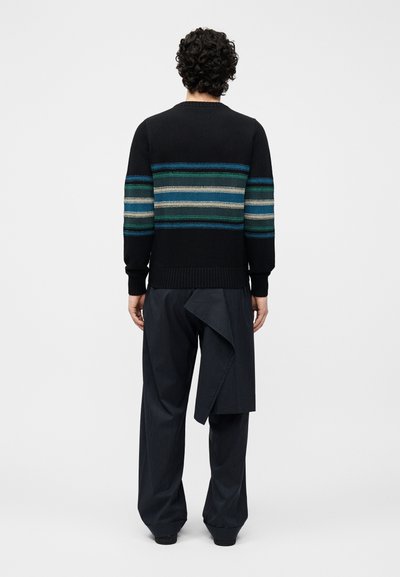Homme aux cheveux bouclés portant un pull noir à rayures horizontales bleues et vertes et un pantalon large à fines rayures avec un détail de tissu drapé, vu de dos.