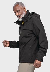 Schwarze wasserdichte Jacke mit Kapuze, ausgestattet mit elastischen Bündchen, durchgehendem Reißverschluss und einer Brusttasche, getragen über einem gelben Shirt und beigen Hosen.