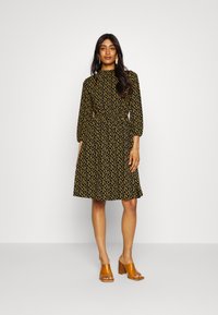 Vestido preto com um padrão de manchas amarelas, mangas longas em balão e cintura ajustada. Combinado com sandálias de salto bloco castanhas.