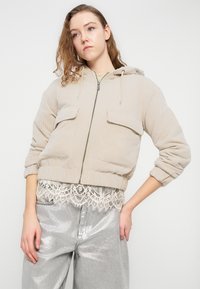 Μπεζ τζάκετ από κορντόρι με κουκούλα, εμπρόσθιο φερμουάρ και δύο τσέπες στο στήθος. Μανσέτες και τελείωμα με ριμπ ύφανση· φορεμένο πάνω από τοπ με δαντελένιο τελείωμα και γυαλιστερό ασημί παντελόνι.